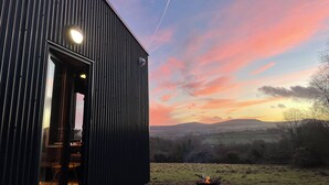 Een modern gebouw met een groot raam en een deur, uitkijkend op een landelijk landschap met een zonsondergang.
