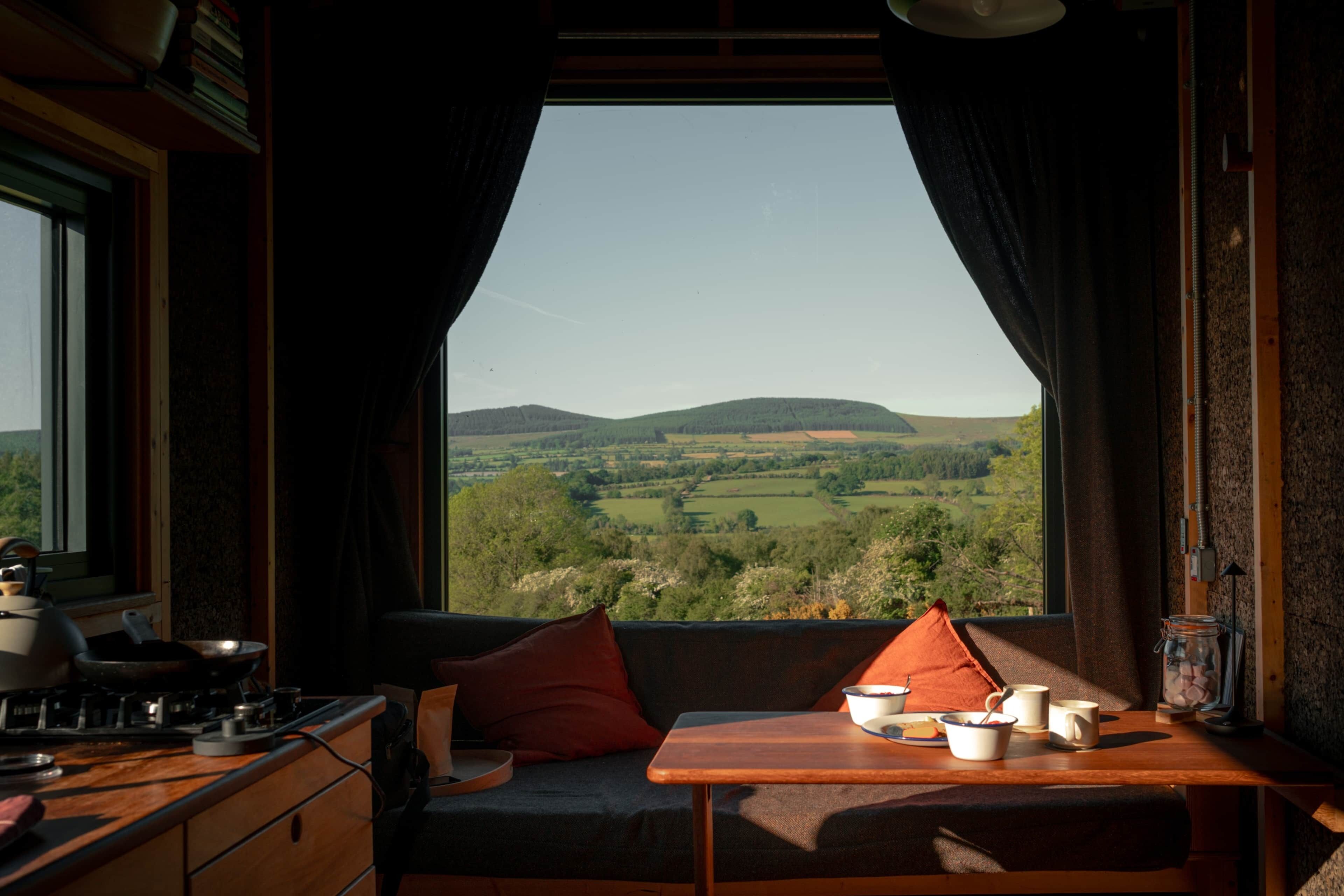 A room with a view of a green landscape, a table with cups and a bowl, and a sofa with cushions.