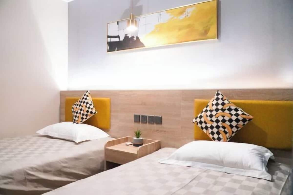 A hotel room with two beds, each with a checkered pillow, a small bedside table with a plant, and a wall-mounted light fixture.