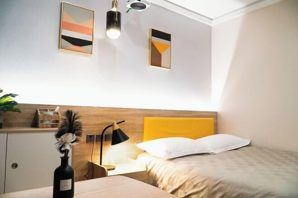 A modern bedroom with a wooden headboard, a bedside table with a lamp, a bed with white bedding, and abstract wall art.