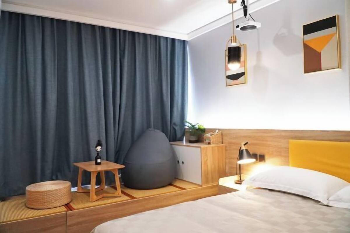 A modern hotel room with a bed, bedside table, lamp, bean bag, and a large window with curtains.