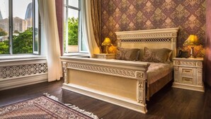 A bedroom with a large bed, ornate headboard, two bedside tables with lamps, a patterned wall, and a view of buildings through a window. - Seven Gates Boutique Hotel (Baku)