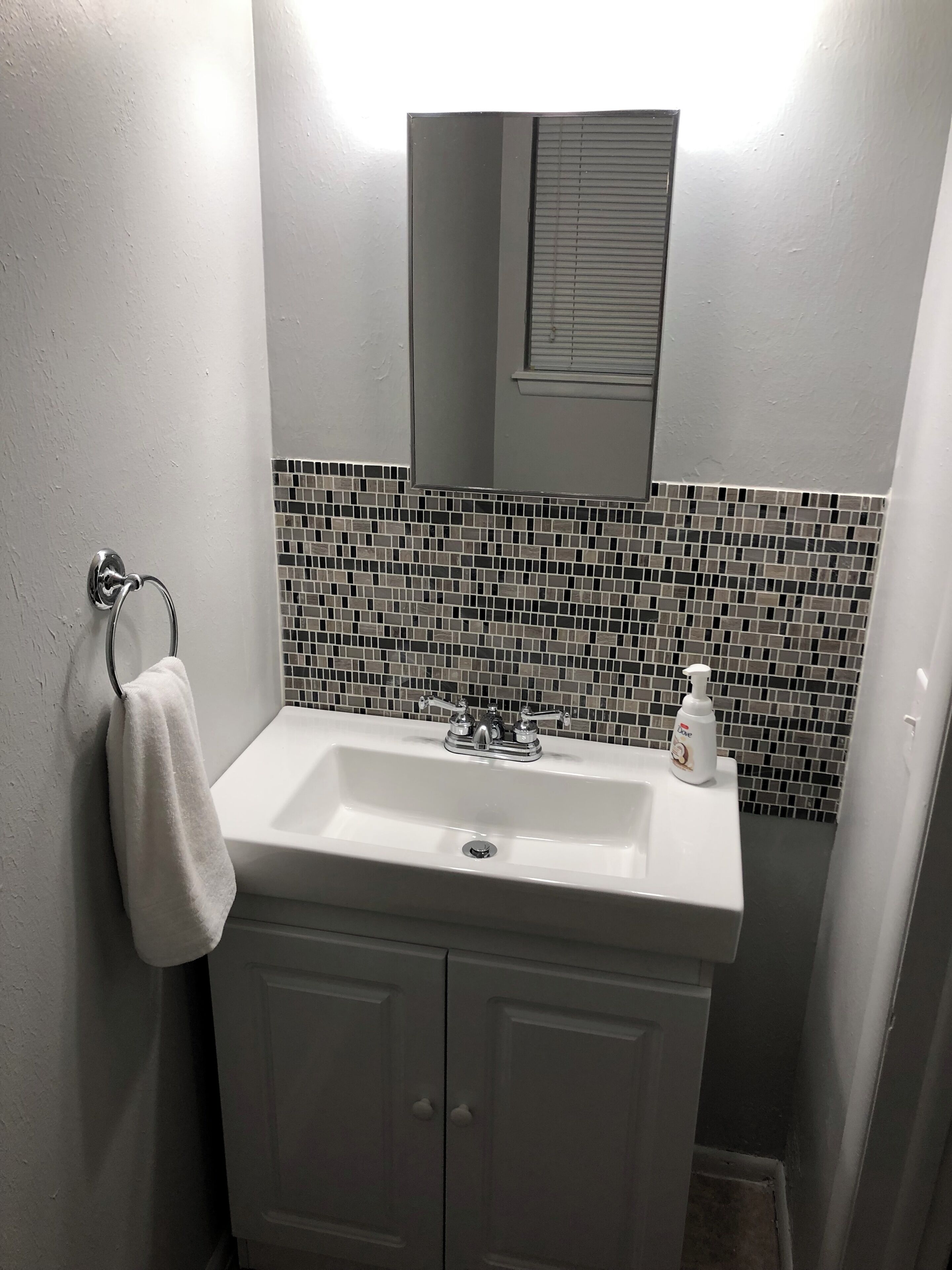 A bathroom with a white sink, a towel hanging on a metal ring, a soap dispenser on the sink, and a tiled backsplash.