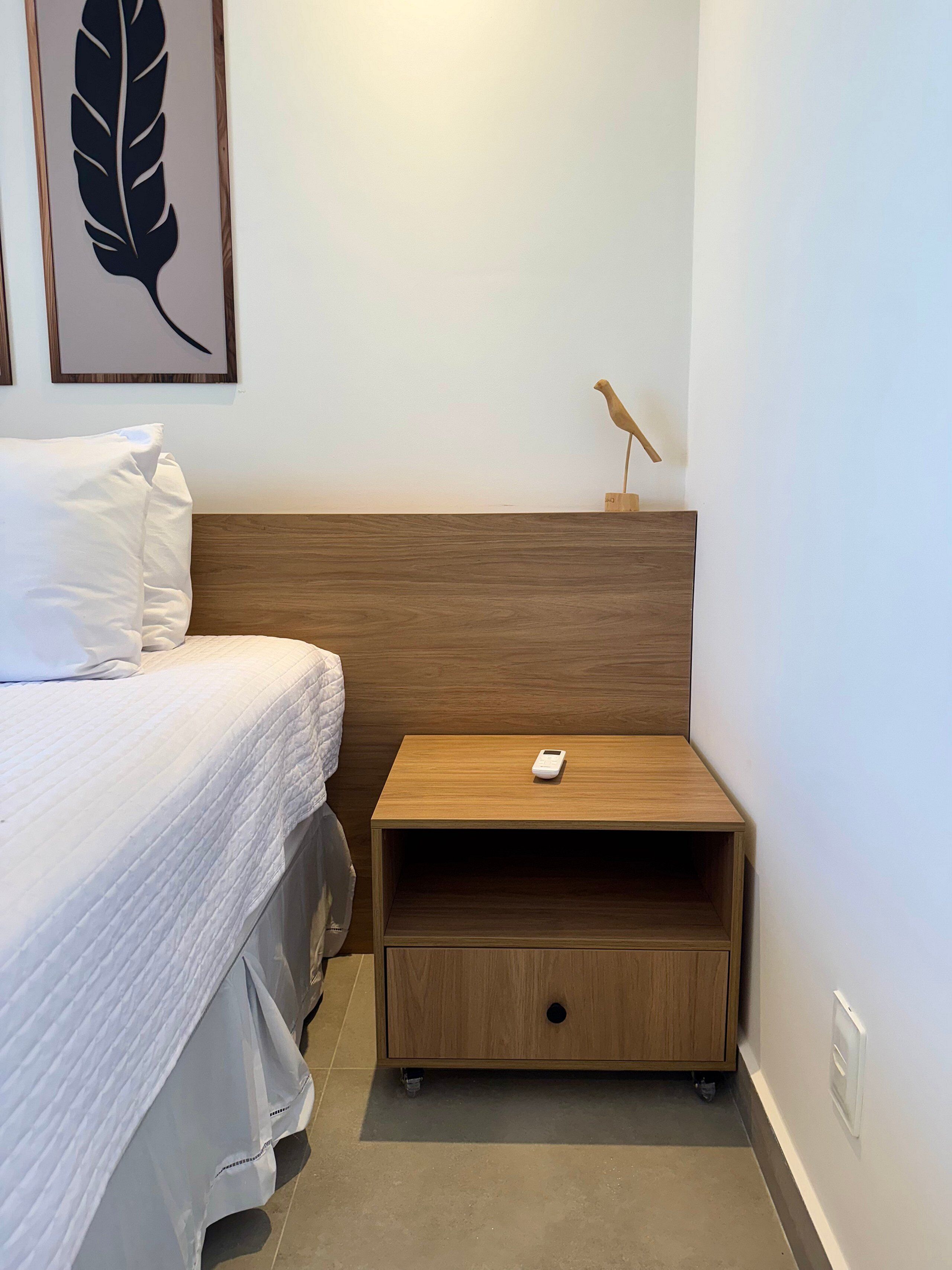 Una cama bien hecha con ropa de cama blanca, una mesita de noche de madera con un teléfono inteligente encima y una escultura de pájaro montada en la pared.