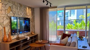 A modern living room with a stone wall, a flat-screen TV, a sofa, a coffee table, and a view of greenery outside. - MHV Milagres - Casa Naluum G4 (Passo de Camaragibe)