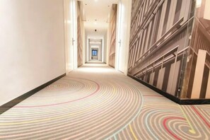 A hotel corridor with a patterned carpet, a view of the lobby, and a door at the end.