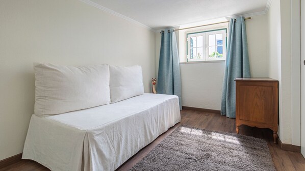 A small, neatly arranged bedroom with a single bed, a wooden nightstand, and a window with curtains.