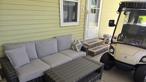 Een modulaire bank met een bijpassende salontafel, een rieten stoel, en een golfkarretje geparkeerd voor een huis met een groene deur en een raam.
