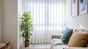 A living room with a bamboo plant, a sofa with cushions, and a window with sheer curtains. - TrindadeFlat w/ AC by LovelyStay (Porto)