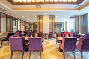 A hotel dining area with multiple tables and chairs, a kitchen in the background, and a modern interior design.