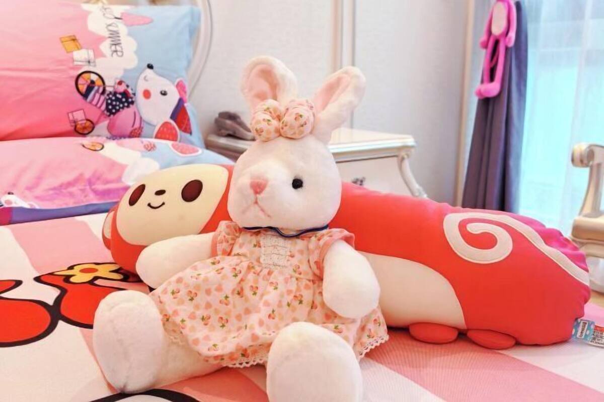 Un lapin en peluche est posé sur un lit, accompagné d’un oreiller à l’effigie d’un personnage de dessin animé et d’un oreiller rouge.