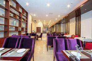 A hotel dining area with purple upholstered chairs, a central aisle, and a long table with a vase of flowers.