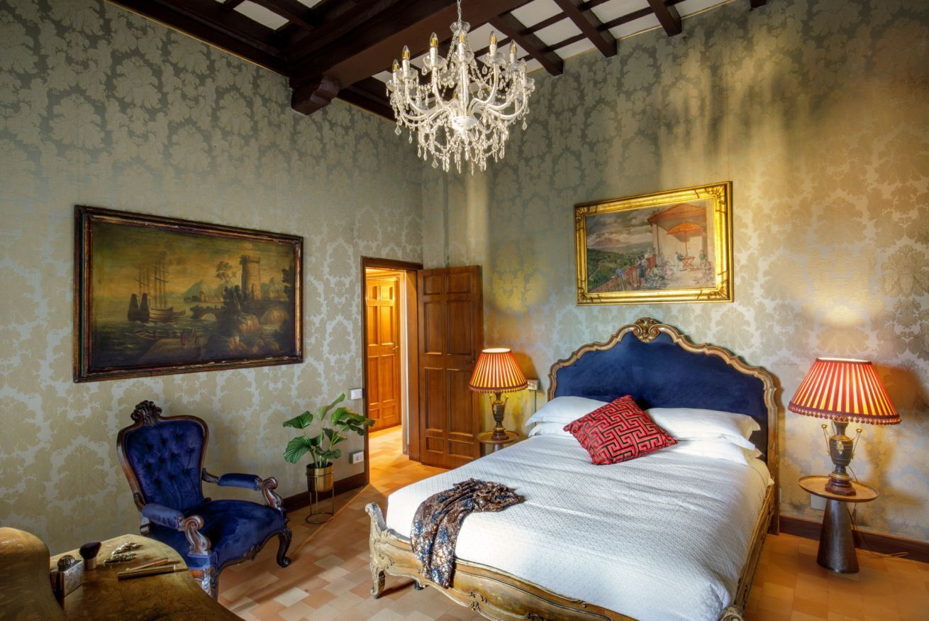 A bedroom with a large bed, a chandelier, a blue armchair, and two lamps.