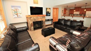 A living room with leather sofas, a stone fireplace, a flat-screen TV, and a kitchen area with wooden cabinets and pendant lights. - Amazing Copper Mountain Views! Steps to Lifts! (Frisco)