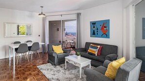 Ein modernes Wohnzimmer mit Essbereich, grauen Sofas, einem weißen Couchtisch und einem großen Fenster mit Blick auf die Stadt.
