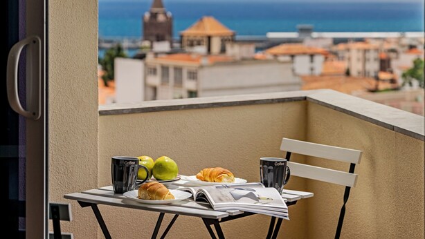 Ein Balkon mit Frühstückstisch, ausgestattet mit Croissants, Äpfeln und Kaffeetassen, mit Blick auf eine Küstenstadt.
