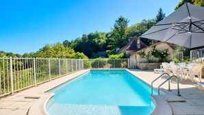 Una piscina rectangular rodeada por una cerca blanca, un muro de piedra con rosales trepadores, y mobiliario exterior con sombrillas.