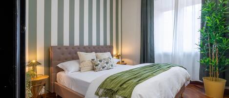 A neatly made bed with a green throw blanket, a tufted headboard, and multiple pillows. There is a potted plant to the right and a lamp on the left side of the bed.