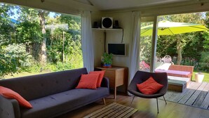 Una sala de estar moderna con un sofá gris, una mesa de centro de madera y un sillón negro con cojines rojos. Hay un televisor sobre un mueble bajo, una planta en maceta y un paraguas verde grande afuera.