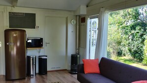 Una sala de estar con un sofá, un banco de madera, un refrigerador, un basurero, un calefactor y una ventana con vistas a la vegetación.
