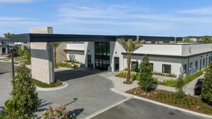 Een modern gebouw met een overdekte entree, aangelegde groenstroken en geparkeerde auto's.