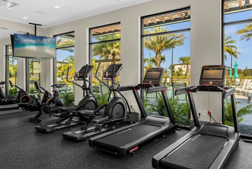 Un gimnasio con cintas de correr, bicicletas estáticas y máquinas elípticas, ventanas amplias con vistas a palmeras y una piscina.