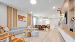 Ein modernes Wohnzimmer mit einer Couch, Couchtisch und Sesseln. Es gibt einen Essbereich mit Tisch und Stühlen. Der Raum verfügt über Holzvertäfelung und einen an der Wand befestigten Fernseher.