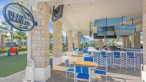 An outdoor patio area with stone pillars, blue and white chairs, and a large TV screen. - Luxe Retreat - Beautiful Modern 7 Bedrooms Home (Davenport)