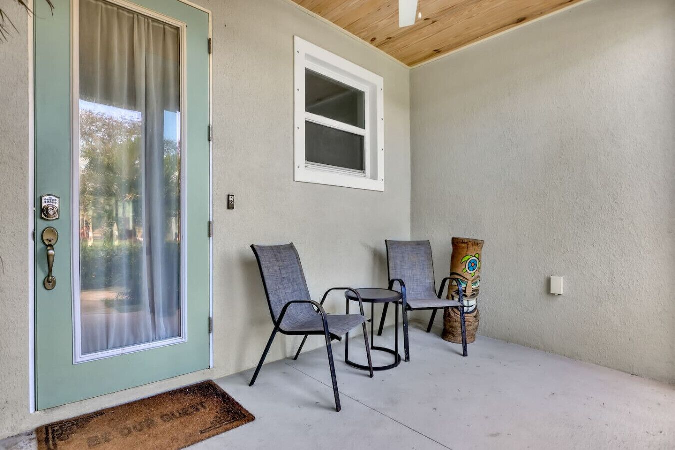A covered porch with two chairs and a decorative pillow, a door with a window, and a small window above the door.