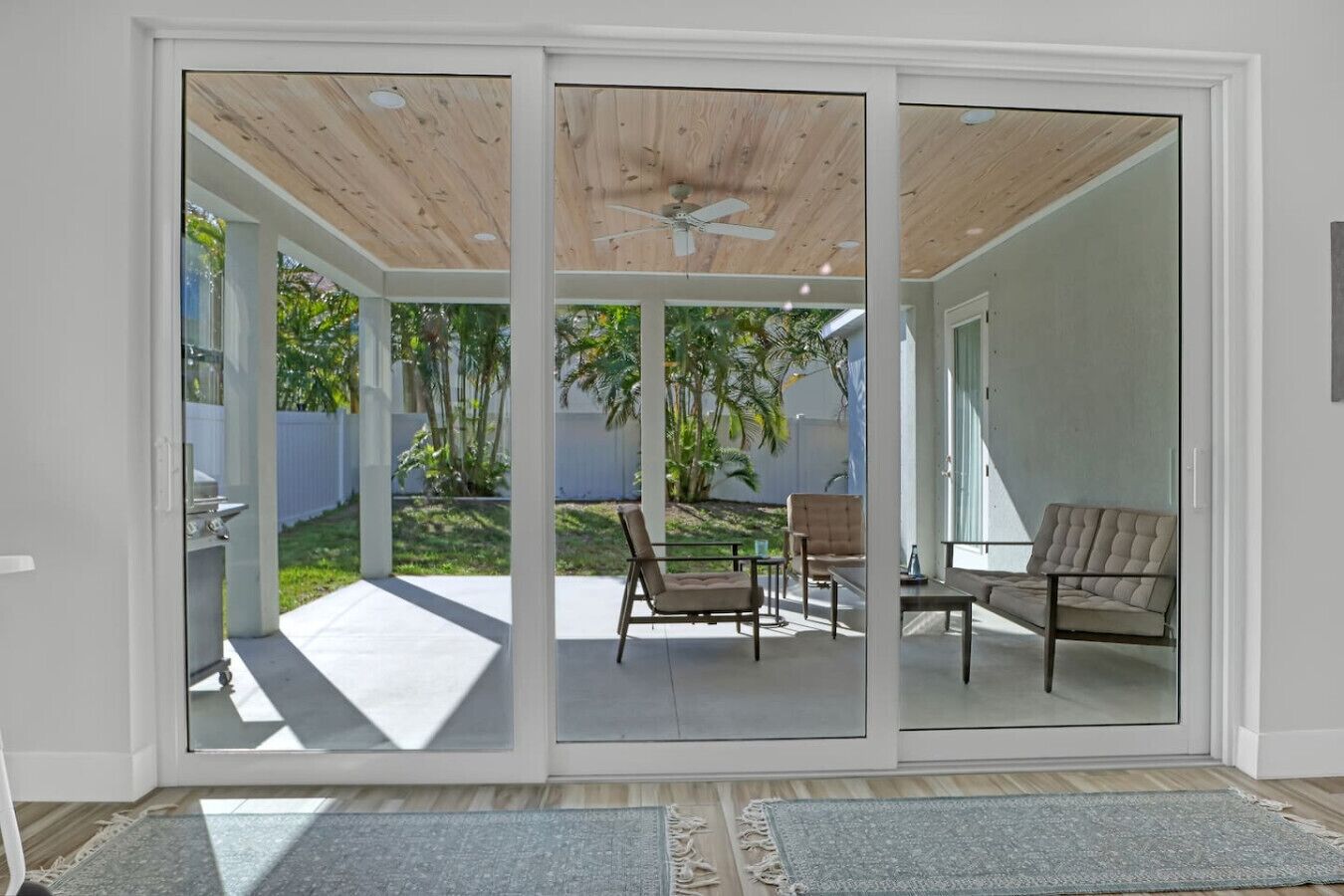Eine moderne Schiebetür führt zu einer Terrasse im Freien, die mit einem Deckenventilator, einer Sitzecke mit zwei Stühlen und einem Tisch sowie einem Garten mit weißem Zaun und viel Grün ausgestattet ist.