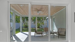 Una porta scorrevole in vetro moderno che dà su un patio esterno con una ventola a soffitto, un'area salotto con due poltrone e un tavolino, e vista su un giardino recintato da una staccionata bianca e con piante.