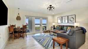A spacious living room with a gray sofa, wooden dining table, and chairs. There is a large window with a view of the outdoors. - The White House on Flagler Beach - Unit 23 (Flagler Beach)