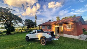 A white SUV parked in front of a brick house with a red roof, a grassy yard, and trees in the background. - Mini Pantanal Paulista with a paradisiacal view, a place to listen to the birds (Ibitiruna)