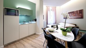 Et moderne køkken med mikrobølgeovn, vask og skabe. Der er et spisebord med stole og en potteplante. Et billede hænger på væggen, og et vindue med gardiner er synligt.