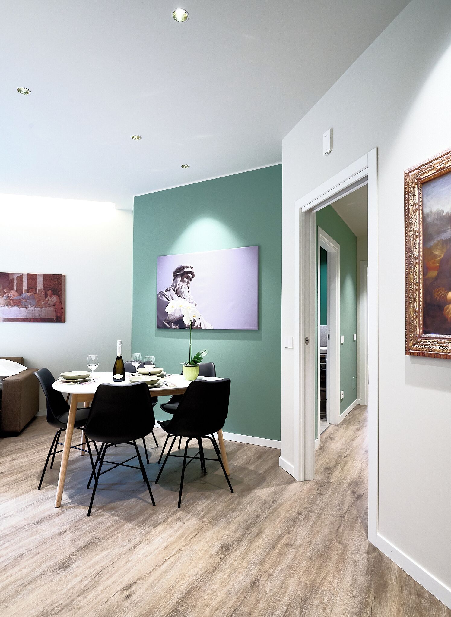 Un comedor con una mesa dispuesta para cuatro personas, una pared verde decorativa y un cuadro sobre la mesa.