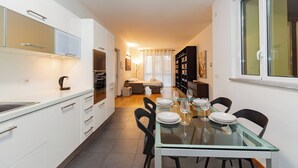 Une cuisine moderne avec des meubles blancs, une table à manger en verre pour quatre personnes et un coin salon confortable avec un canapé et une table basse.