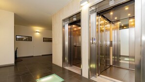 Un ascenseur moderne doté d’un écran numérique, un couloir propre au sol carrelé et un tableau encadré accroché au mur.