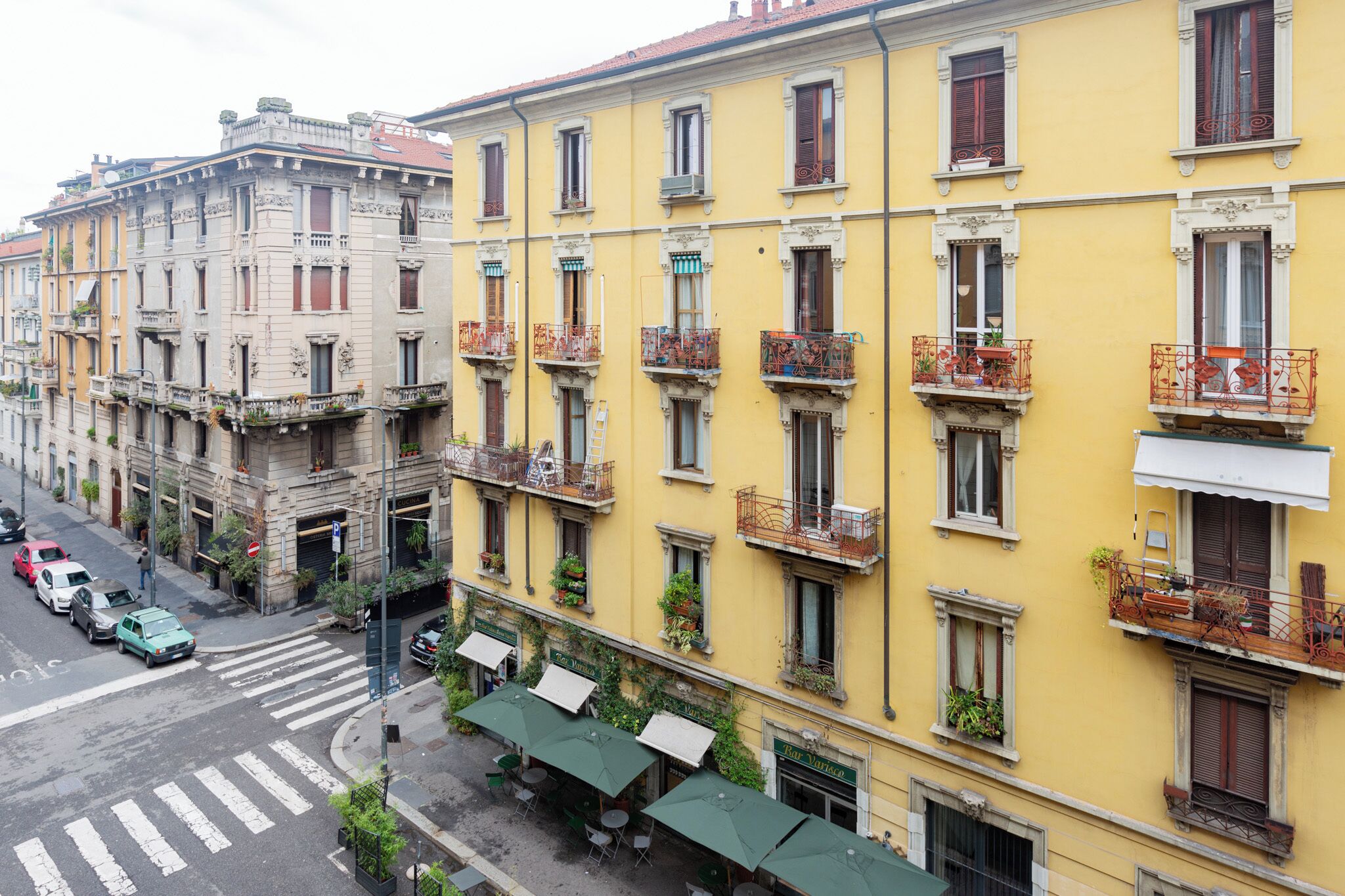 A street view of a row of yellow buildings with balconies, some with plants. There are cars parked on the street and a pedestrian crossing.