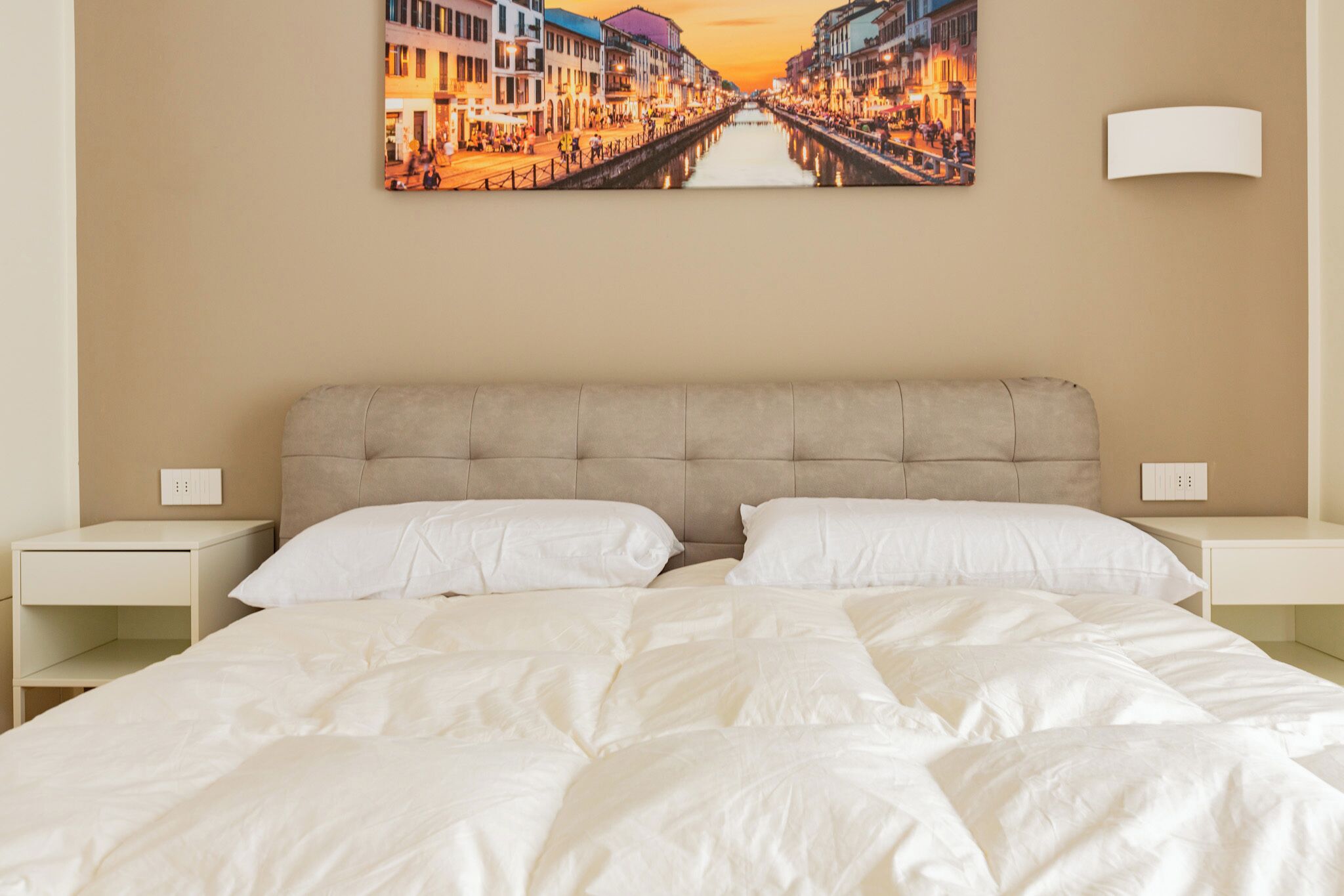 Ein Hotelzimmer mit einem großen Bett, zwei Nachttischen, einem Kopfteil und einem an der Wand hängenden Bild von einer Stadtlandschaft im Abendlicht.