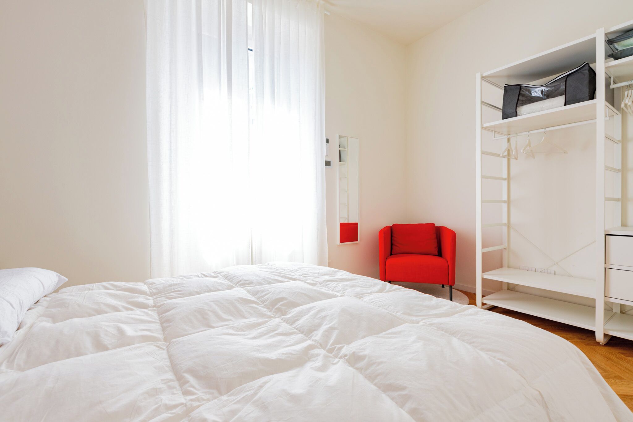 Ein Schlafzimmer mit einem großen Bett, einem roten Sessel, einem weißen Kleiderschrank und einem Fenster mit durchsichtigen Vorhängen.