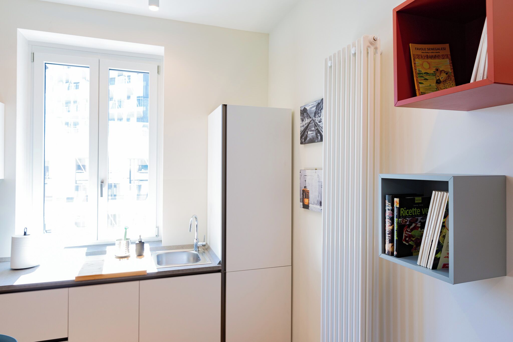 A kitchen with a refrigerator, a sink, and a window. There is a bookshelf with books and a picture frame on the wall.