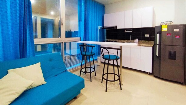 A compact living space with a blue sofa, two bar stools, a kitchen area with white cabinets, and a refrigerator. - a una cuadra de la playa  (Santa Marta)