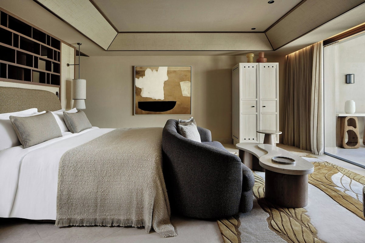 A modern hotel room with a bed, a seating area, a large window, and a cabinet.