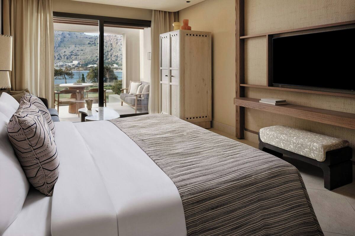 A hotel room with a bed, a TV, a bench, a wardrobe, and a view of a lake and mountains.