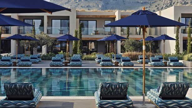 A pool area with lounge chairs and umbrellas, a modern building with large windows, and a mountainous backdrop. - Amoh, A Luxury Collection Resort, Rhodes (Rhodes)