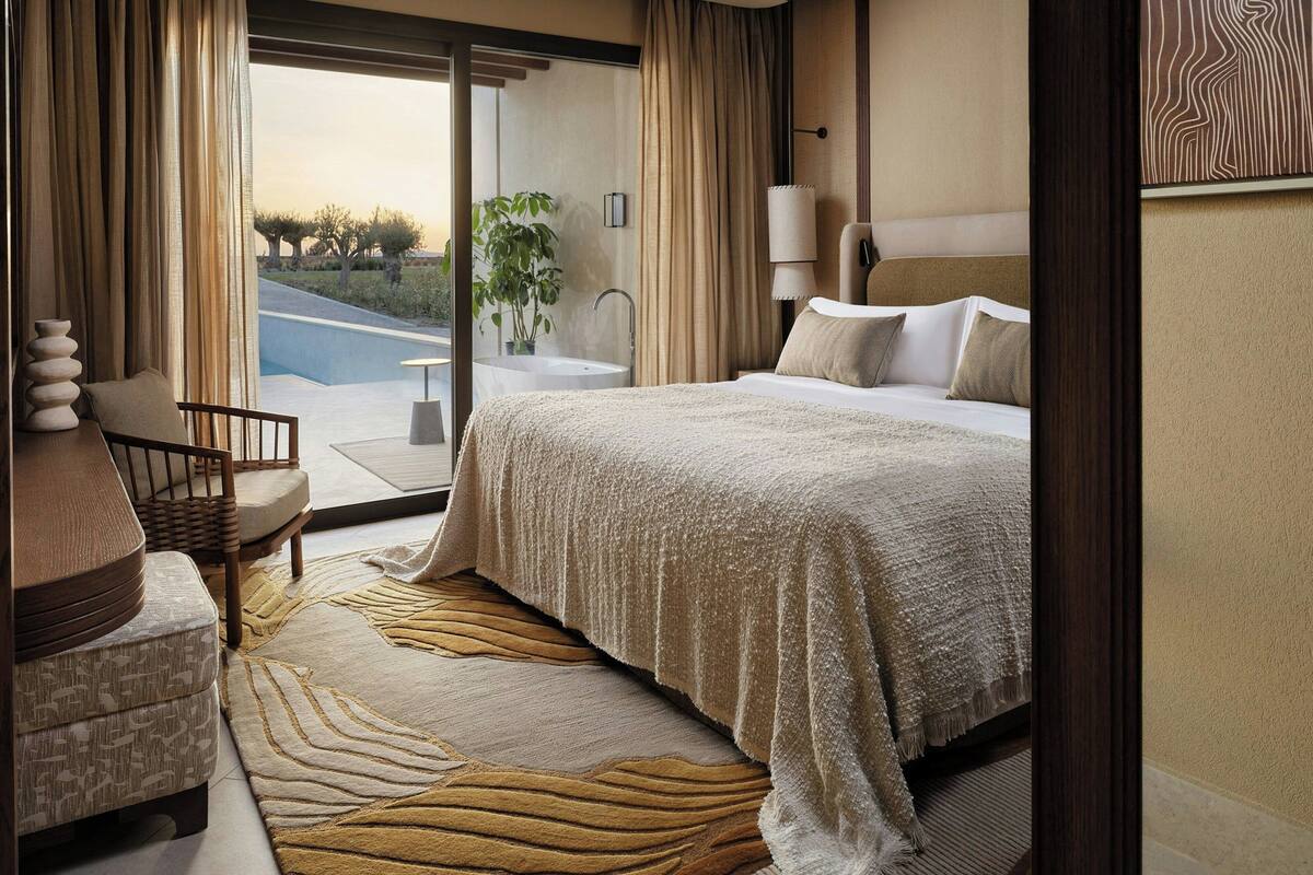 A hotel room with a bed, a chair, a small table, a lamp, a large window, and a view of the outdoors.