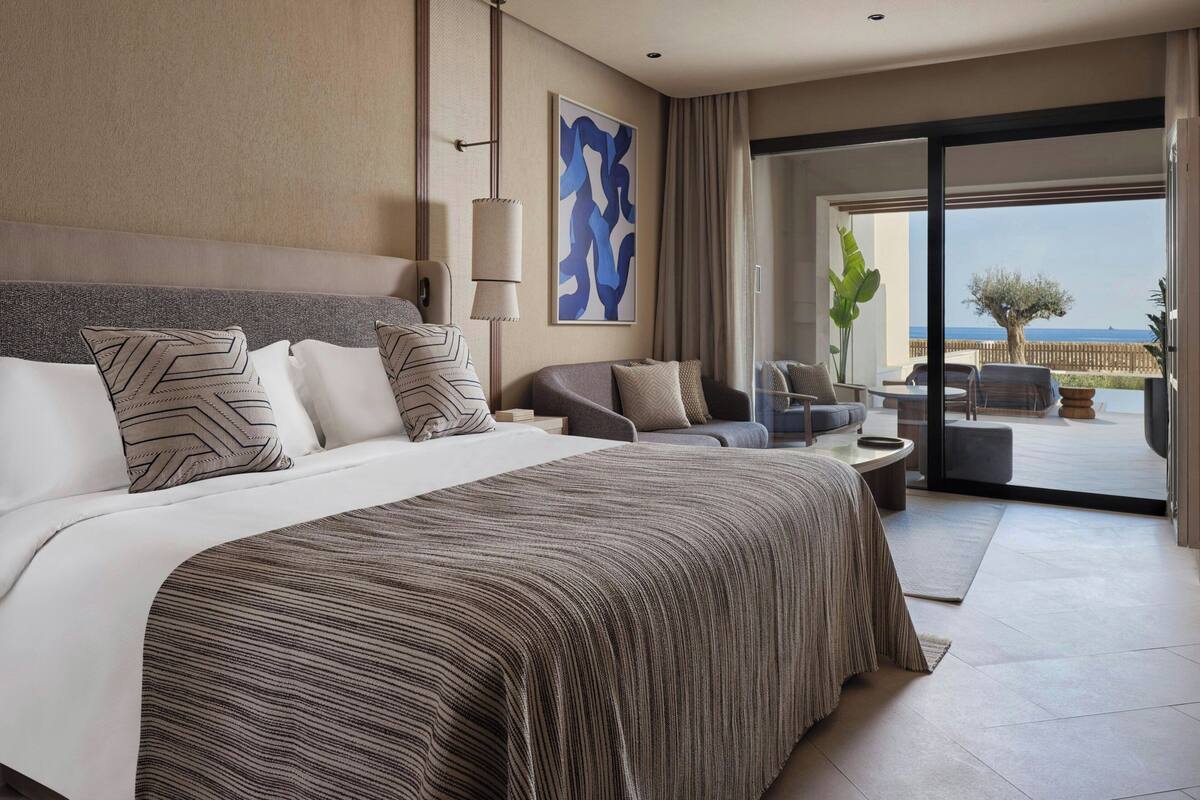 A modern hotel room with a large bed, a sofa, a dining area, and a view of the sea.