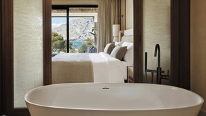 A modern hotel room with a large bathtub, a bed with white linens, and a view of a mountainous landscape through a large window. - Amoh, A Luxury Collection Resort, Rhodes (Rhodes)