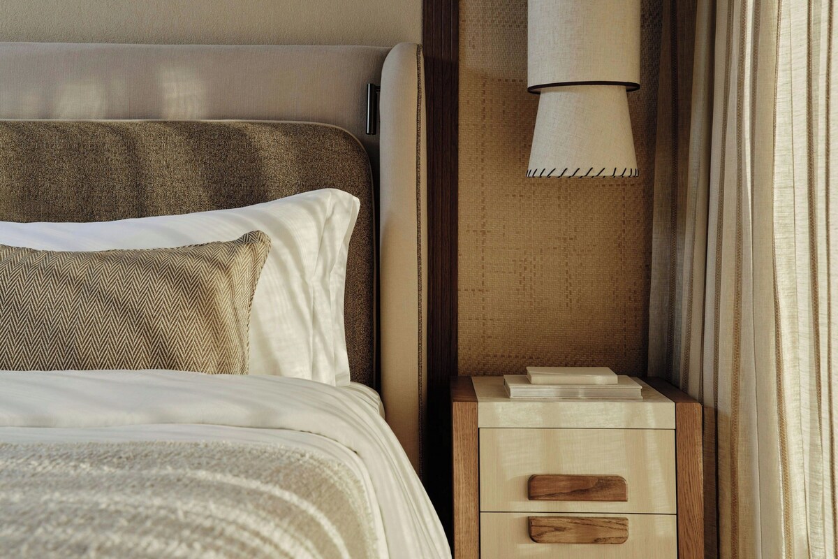 Una cama bien hecha con cabecera, una lámpara de noche y una mesita de noche de madera con cajones.