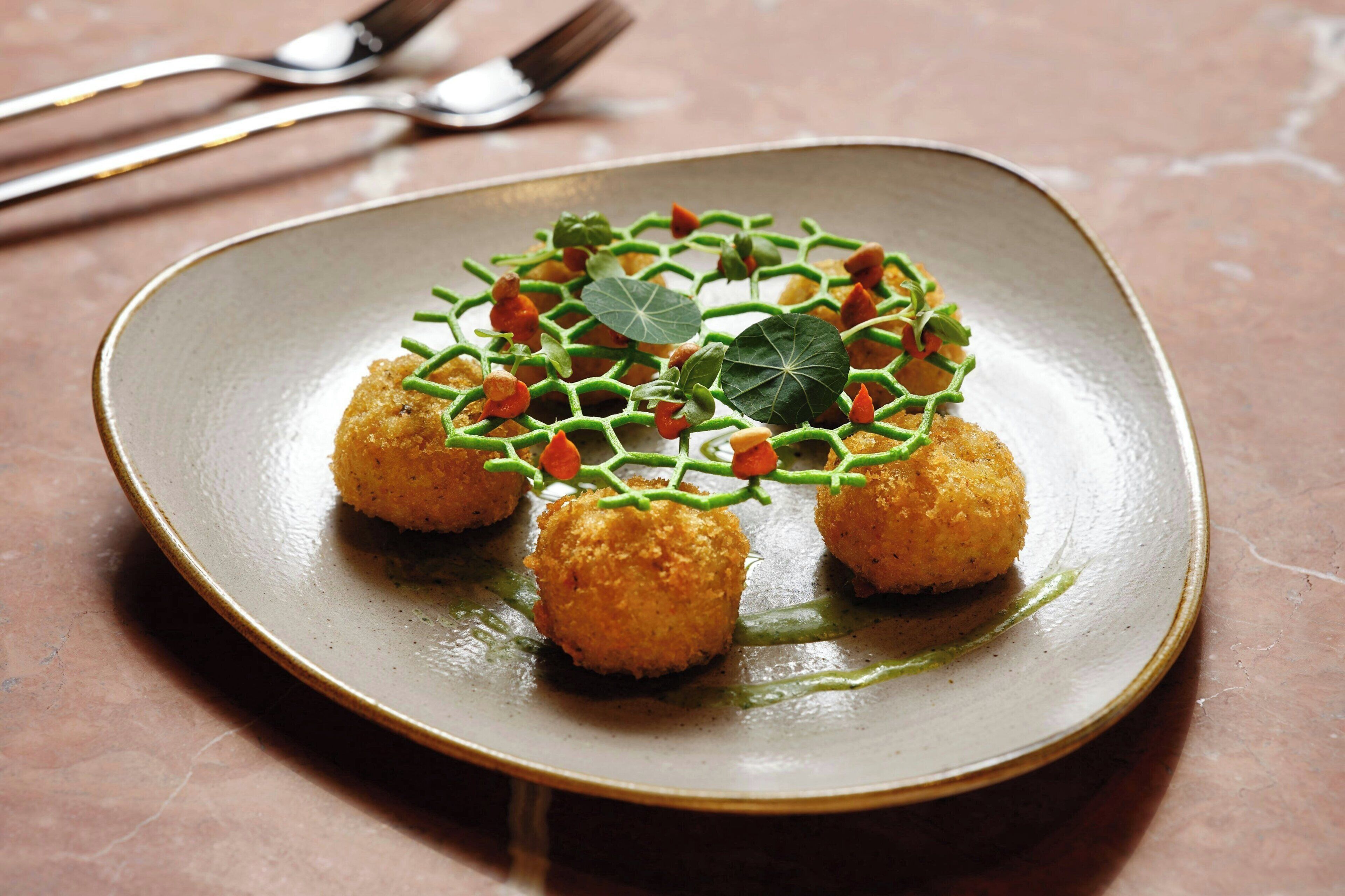 Un plato con tres croquetas doradas, adornado con hierbas verdes y una hoja, sobre un plato de cerámica texturizada.